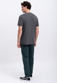Grijze shirt met korte mouwen en ronde hals; groene tapered broek; zwarte sneakers. Subtiele logo-detail op de achterkant van de halslijn.