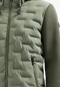 Groene jas met een gevoerde voorkant en een getextureerde gebreide mouwen. Bevat een ritsluiting, een verstelbare capuchon en een zijzak met rits.