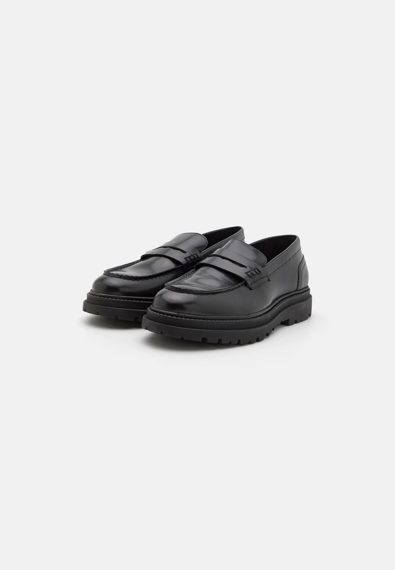 Une paire de mocassins en cuir noir avec des semelles en caoutchouc épaisses et un détail de sangle, présentée sur un fond gris clair uni.