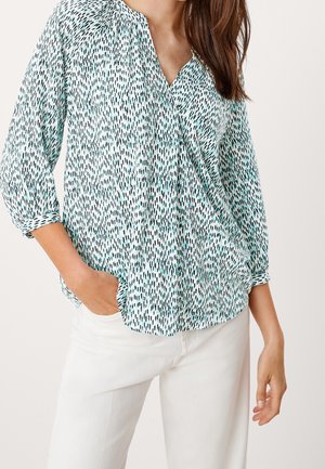 Mujer que lleva una blusa de manga larga con estampado en color azul verdoso y negro con escote en V, combinada con pantalones blancos de talle alto, con la mano en el bolsillo.