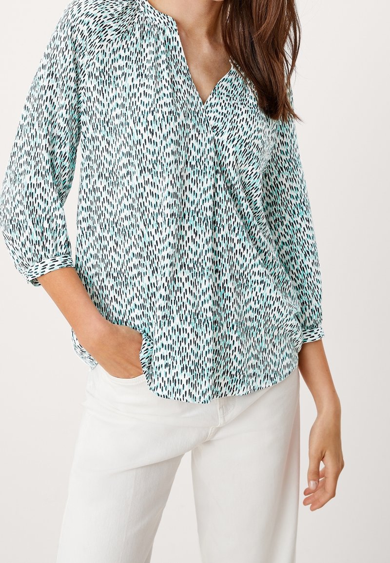 Femme portant un chemisier à manches longues à motif bleu sarcelle et noir avec un col en V, associé à un pantalon blanc taille haute, main dans la poche.