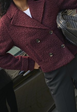 Kobieta w teksturowanej bordowej kurtce dwurzędowej, czarnych spodniach, trzymająca plecioną szarą torebkę, stojąca obok ciemnych drzwi samochodu.