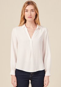 Weiße Bluse mit langen Ärmeln, Kragen, Knopfverschluss vorne und strukturiertem Stoff. Leicht lockere Passform und geraffte Ärmel.
