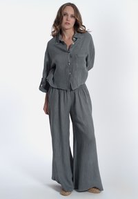 Camicia di lino grigia con bottoni e taschino sul petto, abbinata a pantaloni grigi larghi con cintura elastica e gambe ampie. Indossata con scarpe intrecciate.