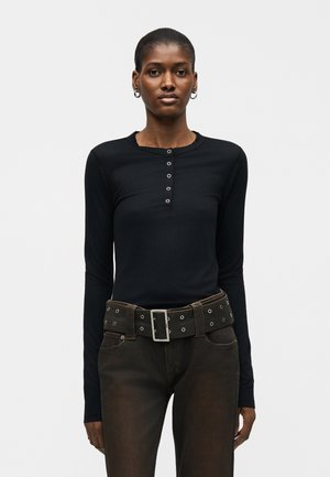 Γυναίκα που φοράει μαύρο πουκάμισο με μακριά μανίκια και κουμπιά, σκούρο καφέ παντελόνι και φαρδιά ζώνη με μεταλλικές τρύπες και μεγάλη ορθογώνια αγκράφα.