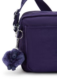 Bolso de tela morado tipo bandolera con un bolsillo en la parte frontal y un llavero de monito de felpa morado a juego adjunto al anillo de la correa.