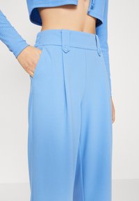 Jasnoniebieskie spodnie o gładkiej fakturze, plisowany fason, zapięcie na dwa guziki i boczne kieszenie. Krótki rękaw o prążkowanym wzorze uzupełnia styl.