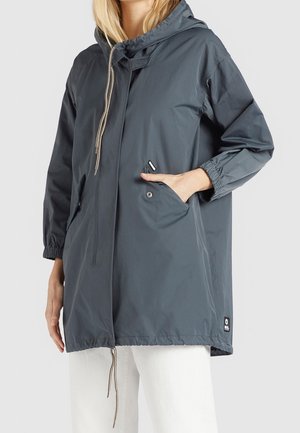 Mujer con una chaqueta holgada de color gris oscuro con capucha, puños elásticos y cordones, una mano en el bolsillo frontal, combinada con pantalones blancos.