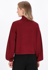 Frau mit blonden Haaren, die einen burgunderfarbenen Rollkragenpullover mit Ballonärmeln und schwarze Hosen trägt, mit dem Rücken zur Kamera gerichtet.