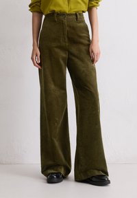 Olijfgroene corduroy wijduitlopende broeken met verticale ribbels, hoge taille en knoopsluiting aan de voorkant, gecombineerd met zwarte schoenen.