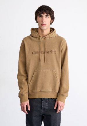 Sudadera marrón con capucha, con un bolsillo frontal y logo bordado. Tejido suave y texturizado con un ajuste relajado, combinada con jeans oscuros.