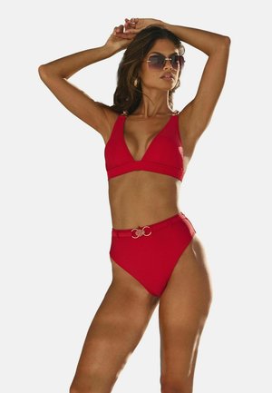 Kobieta w czerwonym bikini z wysokim stanem z paskiem, okularach przeciwsłonecznych i złotych kolczykach kołach, pozująca z podniesionymi ramionami na jednolitym tle.