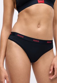 Chiloți negri de tip bikini cu logo-ul roșu "HUGO" pe talie, purtați de o persoană, care arată abdomenul și partea superioară a coapselor.