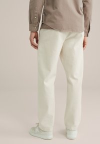 Crèmekleurige katoenen broek met een ontspannen pasvorm, gecombineerd met lichtgrijze sneakers. Het shirt is bruin met knoopmanchetten.