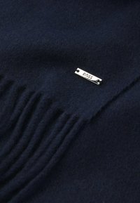 Sciarpa di lana blu scuro con dettagli a frange; presenta un'etichetta in metallo argentato con la scritta "BOSS" incisa. Texture morbida, colore uniforme e finitura liscia.