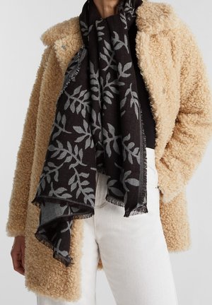 Personne portant un manteau teddy beige, un top noir, un pantalon blanc et une écharpe noire avec un motif de feuilles grises et des bords à franges.