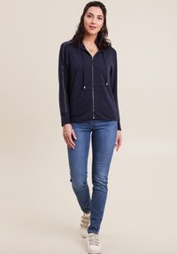 Felpa con zip color navy e cordini, con dettagli argentati lungo le maniche; abbinata a jeans skinny blu e sneakers bianche.