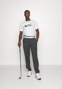 Mann i hvit Nike golf t-skjorte, mørkegrå bukser, hvite joggesko og belte som holder en golfkølle, stående mot en ensfarget hvit bakgrunn.