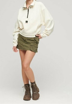 Woman wearing a white quarter-zip pullover, olive green cargo mini skirt, and brown lace-up combat boots, standing with one hand in pocket.
