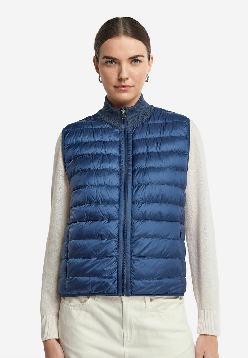 Blauer Steppweste mit hohem Kragen, gestepptem Design und Frontreißverschluss. Getragen über einem cremefarbenen Pullover, der eine glatte Textur und ein leichtes Material aufweist.