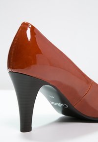 Chaussure à talon haut en cuir verni marron avec un bout pointu et un talon noir texturé. Présente un design sans coutures et une finition brillante.