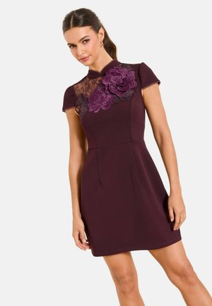 Femeie purtând o rochie mov cu mâneci scurte, detaliu din dantelă pe umăr și o aplicație mare florală mov pe piept.