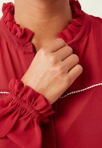 Blusa roja, transparente, con cuello y puños con volantes, que presenta una línea decorativa de pequeñas perlas en la zona del pecho. Textura suave.
