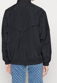 Černá windbreaker bunda s uvolněným střihem, která má strukturovaný povrch, elastické manžety a výrazný design zadního yoku.
