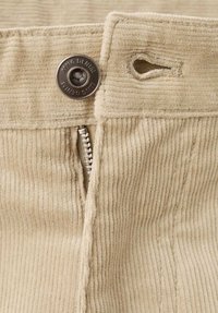 Tela de pana beige con estrías verticales, que cuenta con un botón metálico y cierre de cremallera, además de un pequeño lazo de tela cosido en la pretina.