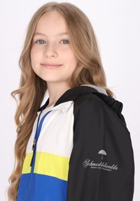 Jachetă colorată cu panouri negre, albe, galbene și albastre. Material ușor, cu închidere tip zipper și glugă. Logo pe mânecă.