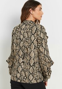 Schlangenmuster-Bluse in Beige und Schwarz. Mit Rüschensleeves, einer glatten Textur und einem taillierten Design. Die Rückansicht hebt die Musterdetails hervor.