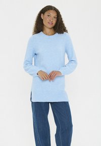 Kvinna med lockigt hår som bär en ljusblå lång tröja och lösa mörkblå jeans, stående framför en vit bakgrund.
