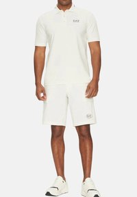 Camisa polo blanca de manga corta, con un sutil logo de EA7. Combinada con shorts blancos, también con la marca EA7, y zapatillas de estilo deportivo.