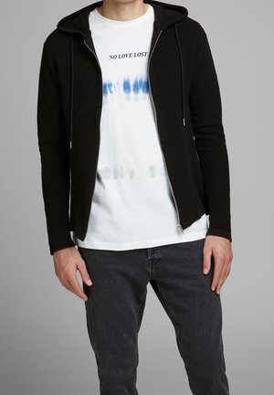 Black zip-up hoodie with ribbed texture, front pockets, and drawstrings, layered over a white tie-dye t-shirt and dark jeans.