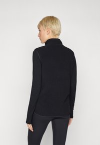 Čierna, textúrovaná fleecová bunda s vysokým golierom, dlhými rukávmi a slim fit strihom, nosená cez čierne legíny, pohľad zozadu.