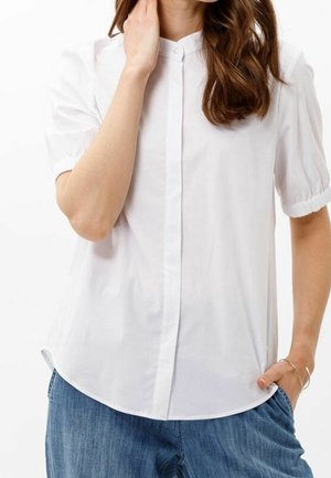 Femme portant une chemise blanche unie à manches courtes boutonnée avec des poignets élastiques et un jean bleu, une main touchant le cou, l'autre dans la poche.