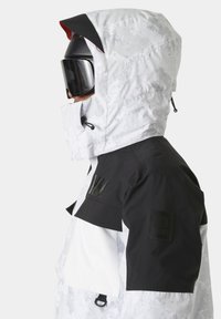 Chaqueta blanca y negra con una superficie texturada, equipada con una capucha, acentos negros y un parche de logo en la manga. Aperturas ajustables visibles.