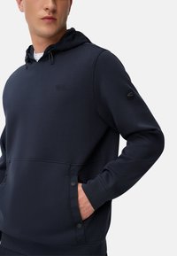 Marineblauwe hoodie met een gladde textuur, voorzak, gemerkt logo en drukknopen als accent op de randen van de zak.