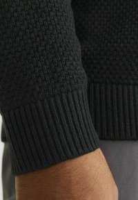 Czarny sweter z dzianiny, z rękawem z fakturowanym wzorem i ściągaczem. Materiał wydaje się miękki i gruby, w jednolitym kolorze.