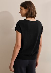 Pull noir à manches courtes, avec un col rond et une finition ajustée. Texture lisse avec une coupe décontractée. Porté sur un pantalon sombre.