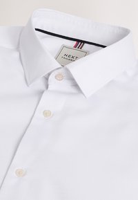 Biela šitá košeľa s rozšíreným golierom, tromi viditeľnými perleťovými gombíkmi a štítkom s nápisom „NEXT Tailoring“ vo vnútri goliera.