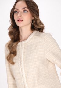 Jachetă din tweed bej cu un model texturat, margine cu franjuri și detalii decorative strălucitoare. Gât rotund și croială ajustată.