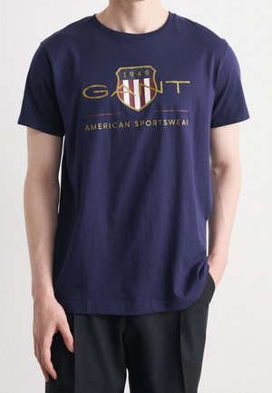 Marineblauwe katoenen T-shirt met een ronde hals, korte mouwen en een gouden schildlogo met rode en witte strepen, gelabeld "GANT AMERICAN SPORTSWEAR."