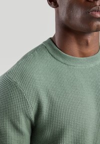 Green knit sweater featuring a textured, waffle pattern and a ribbed crew neckline, shown from the shoulders up.