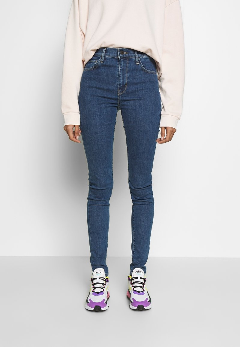 Personne portant un jean skinny taille haute bleu, un haut à manches longues beige clair et des baskets blanches avec des accents violets, noirs et jaunes, se tenant sur un sol gris.
