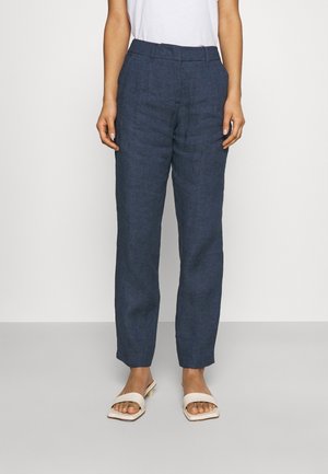 Femme portant un pantalon en lin bleu foncé taillé sur mesure, un top blanc sans manches et des sandales blanches à bouts ouverts, debout devant un fond clair uni.