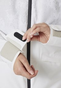 Hvit jakke med teksturert overflate, med borrelås på mansjettene og grå detaljer. Hånd som justerer mansjetten, og viser festet.