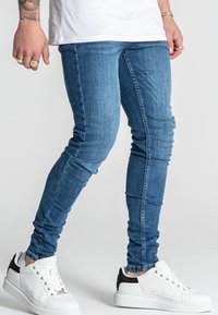 Jeans de denim ajustados en un lavado azul medio con un ligero efecto desgastado; combinados con zapatillas blancas con detalles en negro y una textura caracterizada.