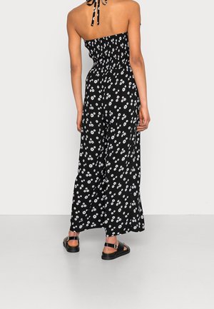 Femme portant une combinaison noire à col halter avec un imprimé floral blanc et des sandales plates noires, debout devant un fond blanc.
