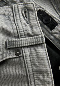 Gros plan d’un tissu en denim gris avec une boutonnière, un bouton noir fixé à un élastique noir et des détails de bouton-pression en métal.